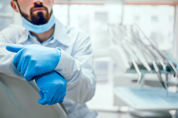 Attractive handsome dentist in protective face mask, white lab coat sits on the chair. dentist equipments on background. Focus on hands.