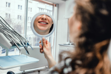 Beautiful young woman looking at mirror with smile in dentist s office.