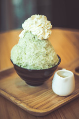 Bingsoo traditional ingredients are, shaved ice, ice cream, in this black cup are green tea bingsoo with whipped cream on top and have condensed milk in a cup ceramic all placed on a wooden tray.