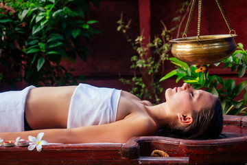 ayurveda massage alternative healing therapy.beautiful caucasian female getting shirodhara treatment lying on a wooden table in India salon © yurakrasil