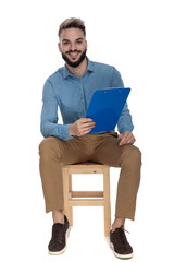 young man sits on wooden chair with clipboard in hands