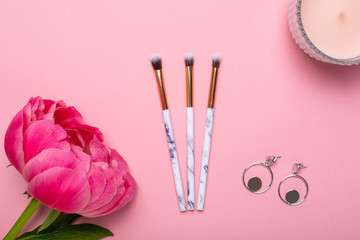 Women's makeup brushes and earrings with a beautiful flower peony on a pink background