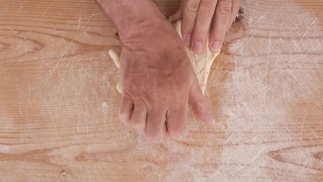 Mani di un uomo stanno impastando la pasta fatta in casa con farina, acqua e sale su un tagliere di legno in cucina.