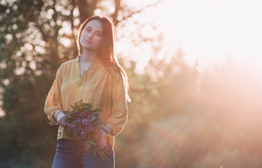 Beautiful young girl, pretty woman in yellow shirt staying in the light of the sun with a bouquet of lilac in her hand. Backlit image, bright, happiness, spring and heat and warmth. Women's Day.