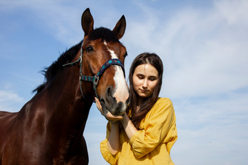 Fototapeta na wymiar Beautiful young girl, pretty european woman in yellow bright shirt hugging adorable brown horse in the sunlight shine with blue sky on the background. Horse farm, horse care, horse riding activity.