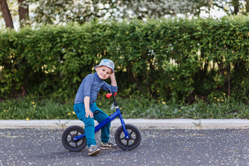 a four-year-old smiling boy in a hat sits on a blue running bike. Bicycle without pedals