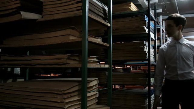 A teenager in a white shirt runs between the shelves with documents in the library. Bookshelves with old literature. Archival room or book depository in the twilight. Concept back to school.