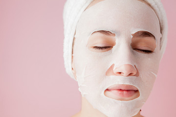 Beautiful young woman is applying a cosmetic tissue mask on a face on a pink background. Healthcare...
