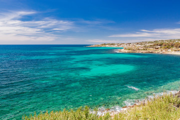 Fototapeta na wymiar Spectacular view of the Apulian coast in Leuca, in Puglia, Salento, Italy. Turquoise sea, clear blue sky, rocks, sun, wind, white clouds, lush vegetation in summer. Populated area with white houses.