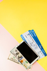 Smartphone, air ticket and dollars on pastel pink and yellow background. Minimal style, flatlay.