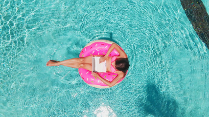 Aerial view of a young brunette woman swimming on an inflatable big donut with a laptop in a...