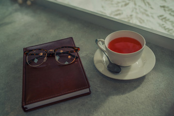 White ceramic tea cup with spoon, notebook near window on stone table, glasses and notebook. Morning planning and inspiration with fruit tea.