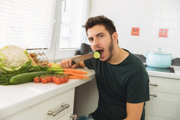 A young handsome man sits in the kitchen and reluctantly eats vegetables. The guy does not want to go on a healthy lifestyle and eat healthy food.