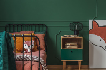 Real photo of a wooden nightstand standing against green wall, between a fox painting and a bed for...