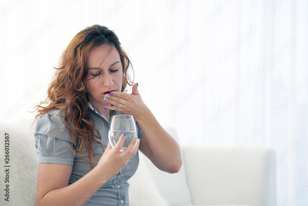 Wall mural Sick woman holding pill glass of water at home on the sofa. Depressed unhealthy woman, about to take antidepressant pill, emergency contraceptive, painkiller for painful periods.