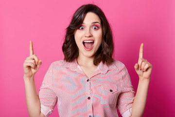 Srudio shot of young brunette girl posing isolated over pink background, points fingers up, having great, female wears striped shirt, has wavy hair, stands with opened mouth. People emotions concept.