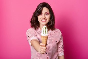 Close up portrait of pleasant looking woman offering ice cream, lady wearing striped shirt stands smiling isolated on pink background, female holding tasty cone. Copy space for advertisment.