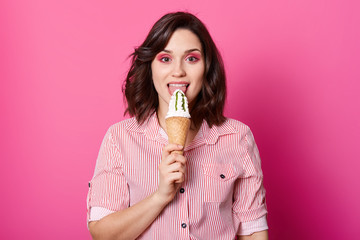 Image of happy woman eating ice cream, has dark wavy hair, wearing stylish shirt with red stripes, has bright makeup, looking at camera with happy expression, isolated on rose studiobackground.