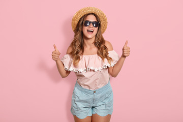 Indoor shot of attractive young happy woman with long hair, wearing stylish clothes, posing with inspired face expression. Active young woman in straw hat having fun indoor, points thumb up.