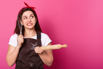 Thoughtful creative worker touching face with whisk, holding rolling pin in one hand, looking...