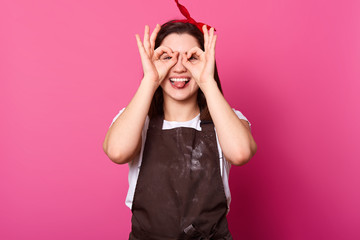 Studio shot of cheerful smiling young woman, covering her eyes with ok signs, expresses joy, model poses against pink background, being glad and joyful. People, body language and gesture concept.