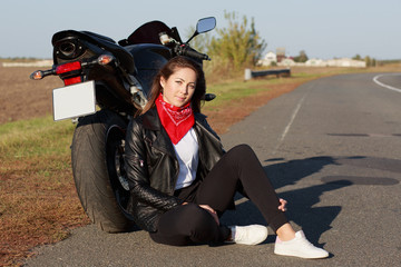 Close up portrait of carefree female motorcyclist wears black clothes and white trainers, sits near...