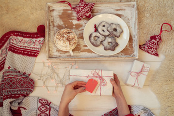 Woman's hands with gift box. Gift with red ribbon, decorations, cup of coffee, cake and red blanket on wooden white floor. Luxury New Year or Christmas gift. Winter time celebration. Toned image.