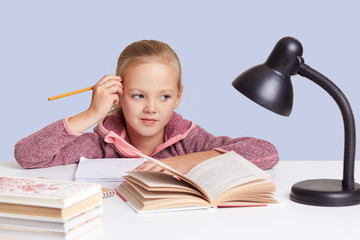 Studio shot of little schoolgirl keeps hand near head, looks with thoughtful expression directly at camera, thinks about homework task, uses reading lamp. Children, education and schooling concept.