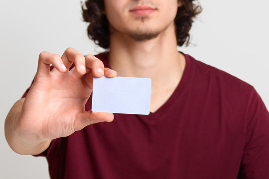 Faceless male stretches hand with blank card for your advertisement or promotion text, poses at white background, man wearing maroon t shirt. Online shopping, purchasing, technology concept.