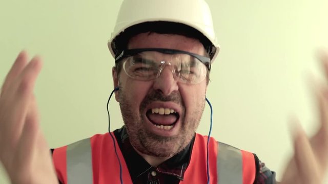 Caucasian unshaven man worker dressed in a reflective vest, protective glasses and white helmet nervously arguing, shouting, waving his arms to the camera. Isolated on light green background