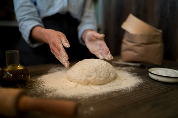 Woman preparing dough at table in bakery for bread or pizza on a rustic wooden surface with natural light. Sprinkling flour over fresh dough on kitchen table. Selective focus. Toned image.