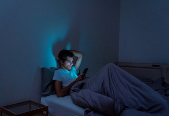 Fototapeta premium Young caucasian sleepless man bored in bed surfing on the Internet addicted to mobile phone