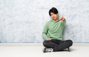 Asian man sitting on the floor showing thumb down with negative expression