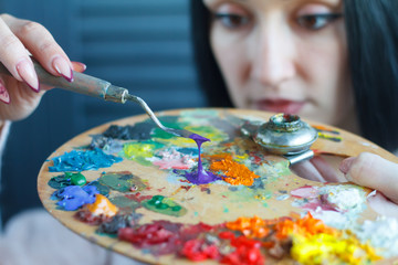 Closeup of a young woman with black hair mixes paint on a palette with a spatula against a white canvas, creating an oil painting