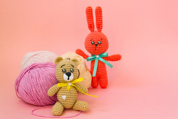 A brown knitted bear sits on a pink background, behind it is a ball of pink and yellow yarn, and an...