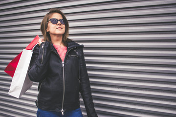 Content young woman wearing sunglasses and a leather jacket is holding her shopping bags over her shoulder – Pretty lady in a fashionable outfit looking away and carrying bags