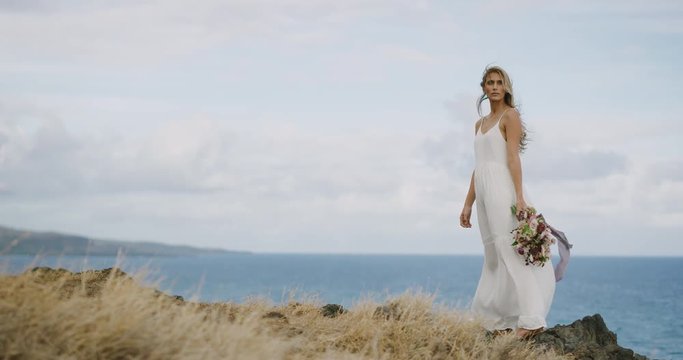 Attractive young woman in a white dress holding a flower bouquet, beautiful woman standing on a golden bluff by the ocean with hair blowing in the wind, bohemian bride lifestyle