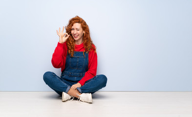 Redhead woman with overalls sitting on the floor showing ok sign with fingers