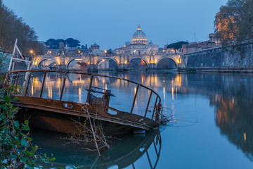 River Tiber in Rome and St Peters Cathedral at night. In the foreground a sunken rusty ship on the shore. Buildings are illuminated. Shore with trees and reflection of the illuminated buildings