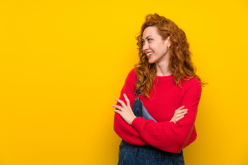 Redhead woman with overalls over isolated yellow wall standing and looking to the side