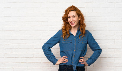 Redhead woman over white brick wall posing with arms at hip and smiling