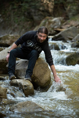 Male standing on a rock in the middle of a water near a waterfall with his hand in the river
