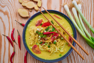 Top view on Soto ayam, spicy indonesian or malaysian soup consist from chicken meat, rice noodles, curry spice, spring onion,tomatoes and coconut milk served in bowl on the wooden table with chilies.