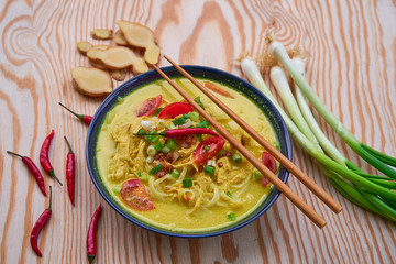 Close up picture of Soto ayam, spicy indonesian or malaysian soup consist from chicken meat, rice noodles, curry spice, spring onion,tomatoes and coconut milk served in bowl on the wooden table.
