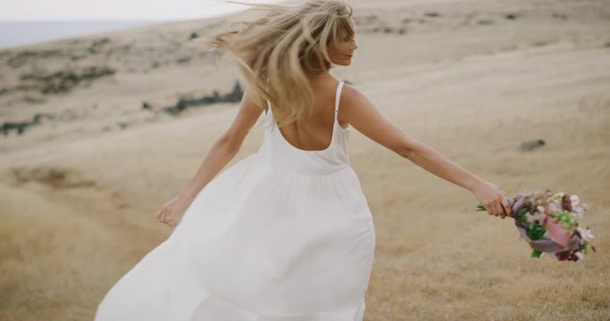 Beautiful young woman in a white dress smiling and twirling in a golden field holding a flower bouquet with her hair blowing in the wind in slow motion, bohemian bride lifestyle