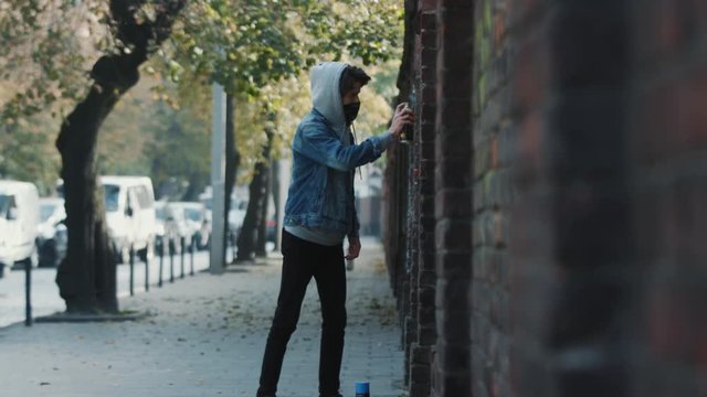 Young male street artist drawing graffiti with aerosol spray on the brick wall in modern town. Urban art.