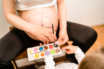 Little blonde girl painting little baby brother or sister on her mothers pregnant belly with water colors and paint brush