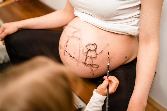 Little blonde girl painting little baby brother or sister on her mothers pregnant belly with water colors and paint brush
