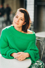 Outdoor fashion portrait of beautiful woman with dark hair, wearing green pullover, posing in street cafe