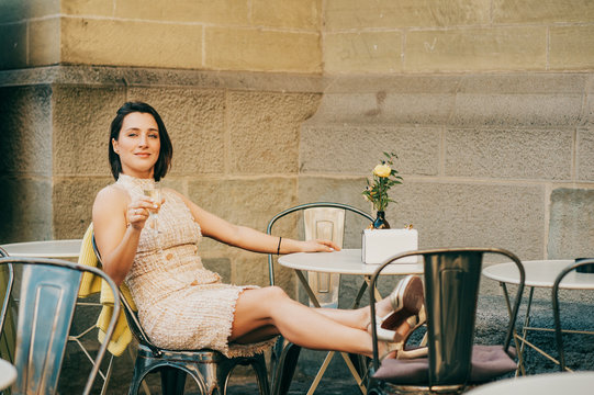Beautiful stylish woman resting in the street cafe with glass of wine, legs on the chair, wearing fashionable dress